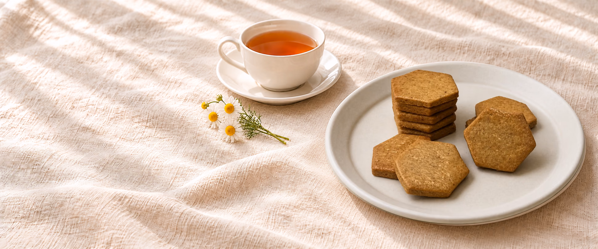 Sablés Rêve Tonka sur leur assiette ivoire, tasse de thé sur sa soucoupe séparée et petites fleurs de camomille sur une nappe en lin