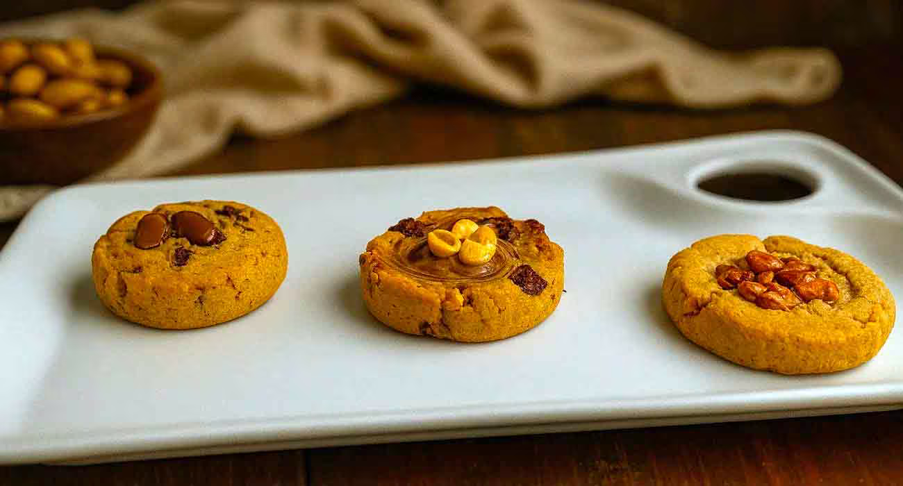 Trois cookies artisanaux aux garnitures gourmandes : chocolat, praliné noisette et fruits secs, présentés sur plateau blanc